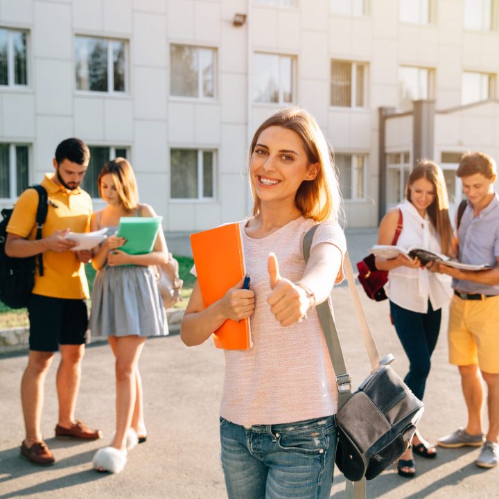 Happy beautiful girl standing with note books and backpack showing thumb up and smiling, standing near university building, her friends are behind